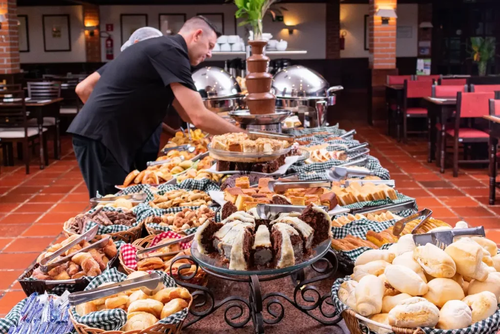 Mesa de buffet completa com pães caseiros, bolos, frutas e cascata de chocolate no Hotel Colonial.