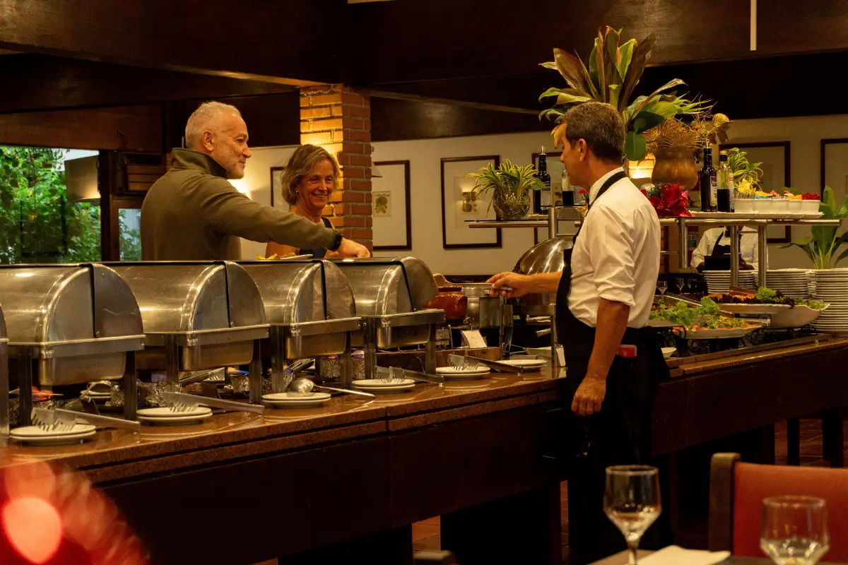 Equipe do Hotel Colonial servindo no buffet de pratos quentes com variedade e higiene.
