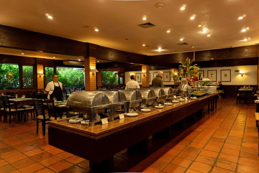Estação central de buffet quente no restaurante do Hotel Colonial Iguaçu, mostrando o amplo salão, janelas com vista verde e equipe pronta para servir.