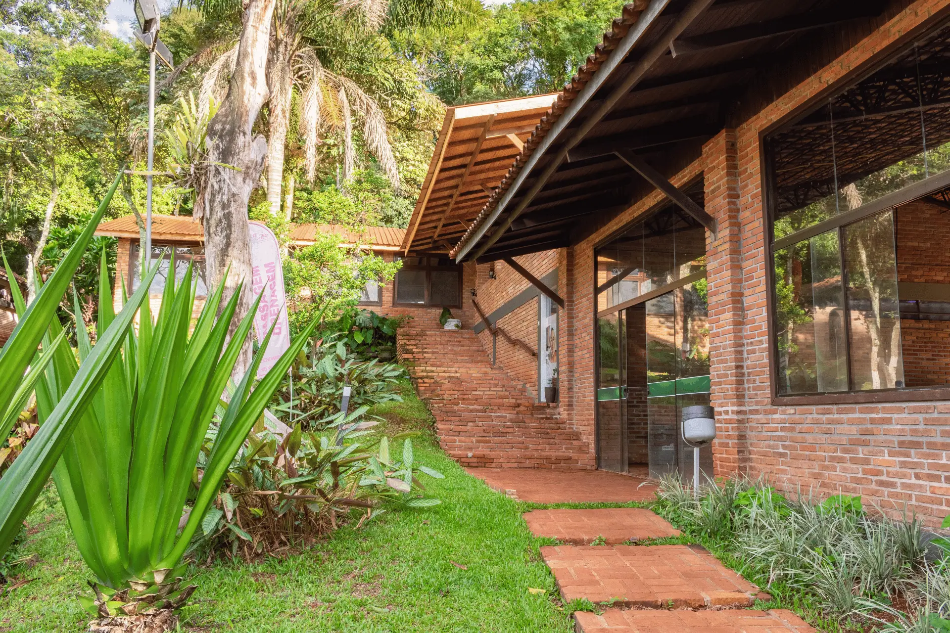 Fachada rústica de tijolos do Hotel Colonial cercada por jardins e natureza, com acesso ao espaço de estética e serviços de massagem e drenagem.