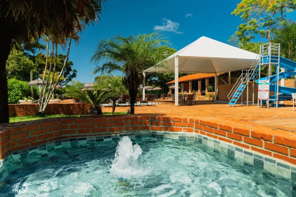 Área de lazer externa do Hotel Colonial Iguaçu com piscina infantil, fonte d'água e tobogã azul ao fundo, cercado por palmeiras e natureza.