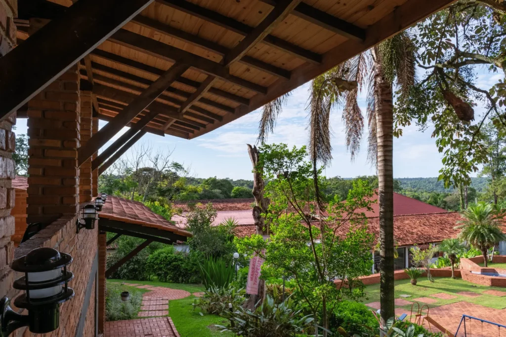 Vista da varanda com colunas de tijolos aparentes, lanternas decorativas e teto de madeira, olhando para os jardins e palmeiras do Hotel Colonial.