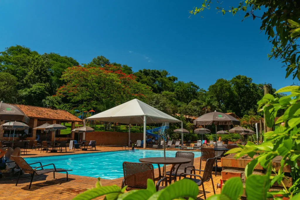 Ampla piscina externa com águas azuis, cercada por espreguiçadeiras, mesas com guarda-sóis e muita natureza no Hotel Colonial Iguaçu.