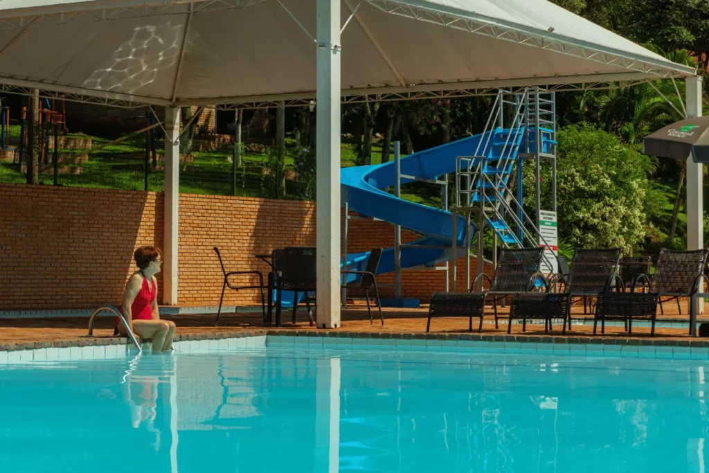 Hóspede relaxando na borda da piscina do Hotel Colonial, com tobogã azul e área de lazer coberta ao fundo em um dia de sol.