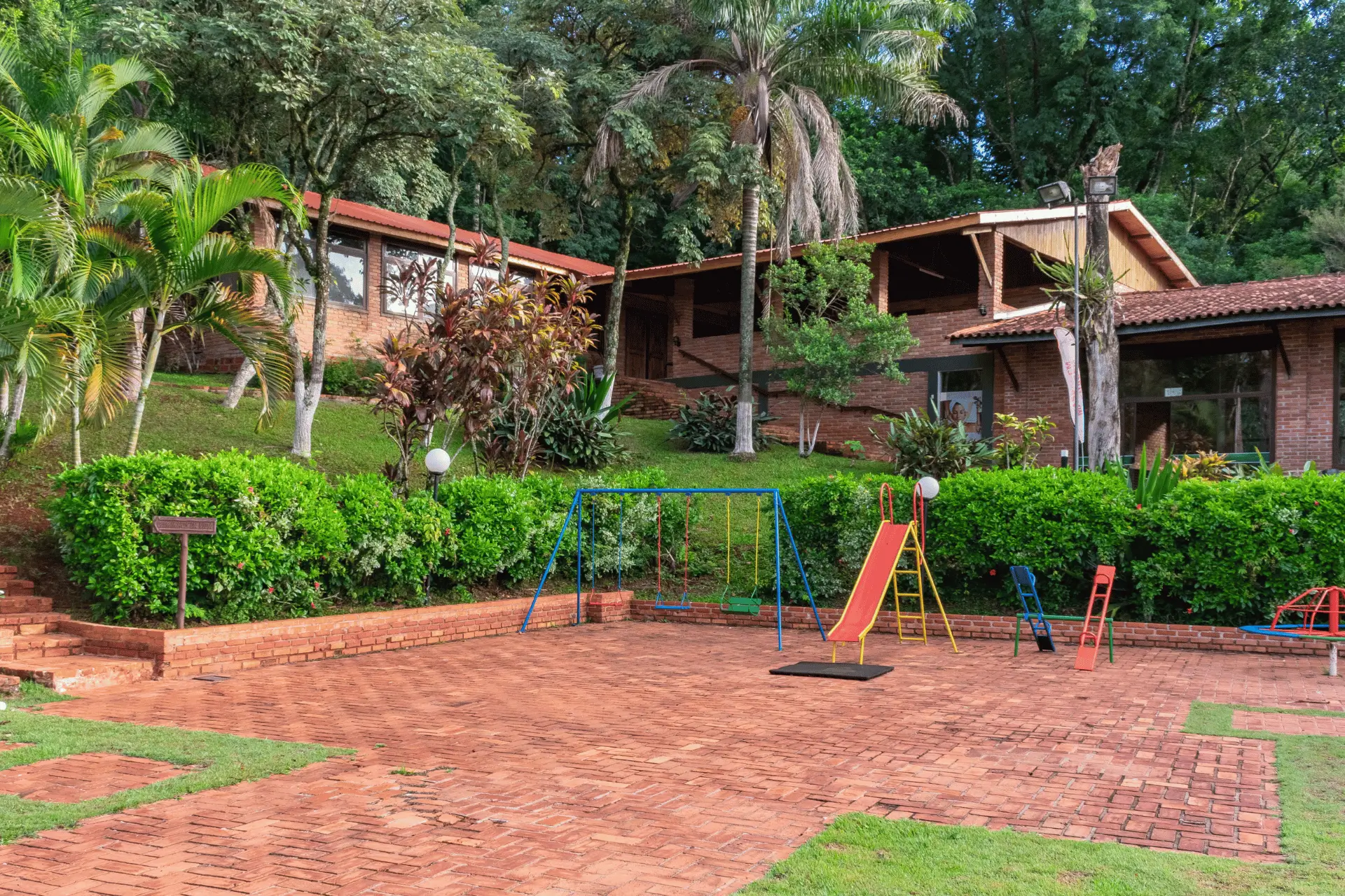 Parquinho infantil externo no Hotel Colonial com balanços, escorregador e gira-gira sobre piso de tijolos, cercado por jardins e natureza.