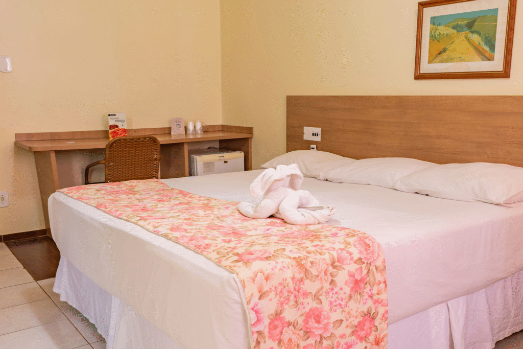 Quarto do Hotel Colonial Iguaçu mostrando cama com decoração de toalha, bancada de trabalho com cadeira, frigobar e menu de serviço de quarto.