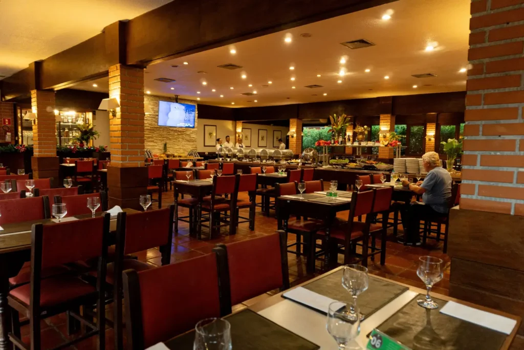 Vista geral do salão do Restaurante Villa Colonial, com mesas postas, cadeiras de madeira, colunas de tijolos rústicos e buffet ao fundo com TV.
